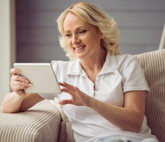 Beautiful old woman Beautiful old woman in casual clothes is using a tablet and smiling while sitting in armchair at home