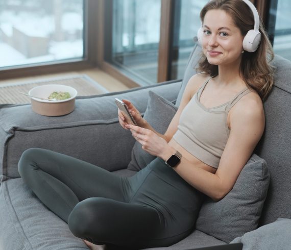 Happy girl in sportswear with headphones and telephone on sofa Young and joyful girl in sportswear rests after workout sitting on sofa with headphones and telephone in apartment on skyscraper.