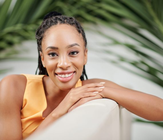 Pretty young Black woman sitting on armchair with palm leaves behid and smiling at camera